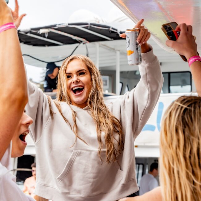 A woman in a beige sweatshirt and swimsuit bottom is joyfully dancing and holding a drink can on a yacht. She is surrounded by others from the social club who are also celebrating, with another boat visible in the background.