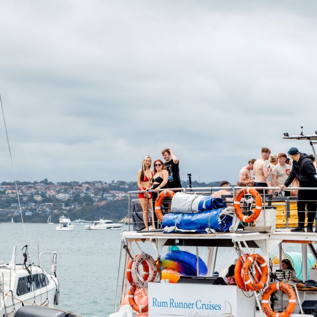 A group from the Social Club is on the "Rum Runner Cruises" yacht, enjoying a day out on the water. Some stand while others lounge on deck. The sky is cloudy, and other boats dot the background.