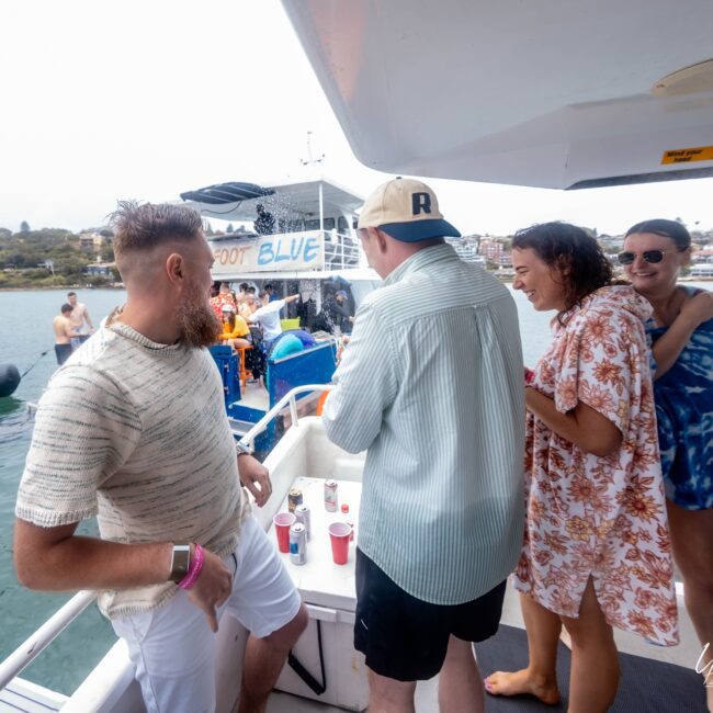 A group of people on a yacht enjoy a sunny day. Three individuals stand by a railing with drinks, smiling and laughing. Others are on a nearby floating platform. The backdrop is a cityscape across the water, creating the perfect setting for this lively social club gathering.