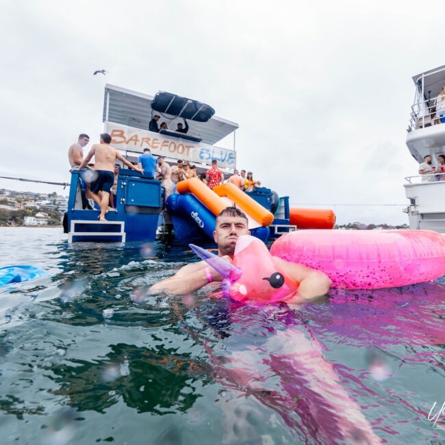 A man in a pink unicorn floaty drifts near a boat with a "Barefoot Blue" sign, embodying the lively spirit of the Yacht Social Club. Other revelers on the boat enjoy inflatables and slides, while hillside homes peek through the cloudy sky.