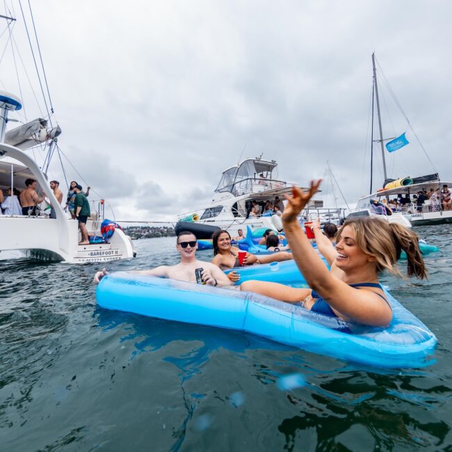 Two people relax on inflatable floats in the ocean, surrounded by a social club of anchored yachts where people are joyfully socializing on board. The sky is overcast, and the mood is lively and festive.