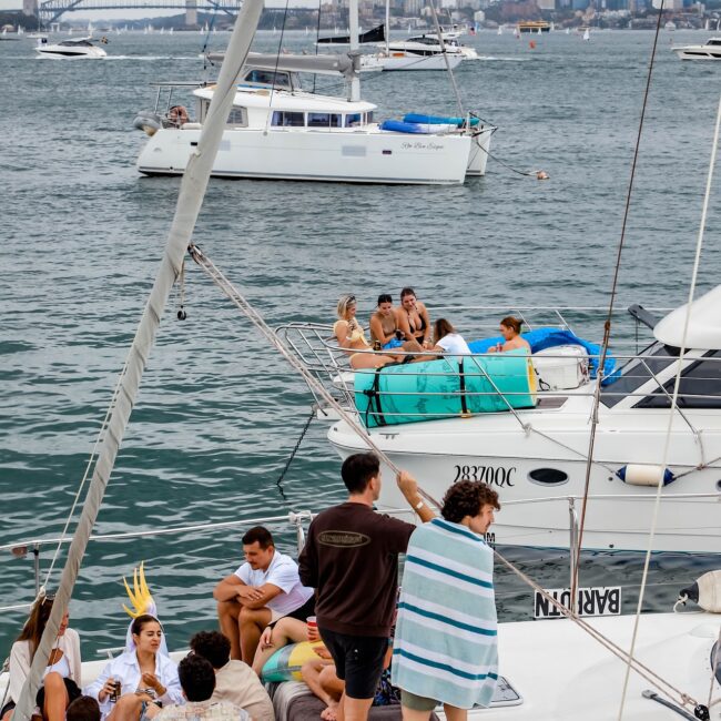 On a cloudy day, members of the social club unwind on a yacht. Some sit while others stand, wrapped in towels, enjoying the serene backdrop of boats on the water and a city skyline with a distant bridge.