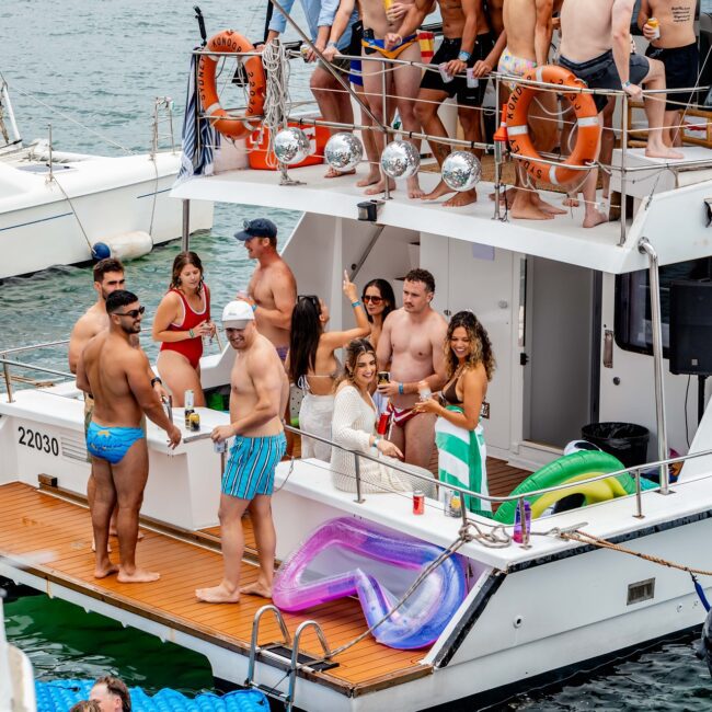 A vibrant Yacht Social unfolds as a lively group in swimsuits revels in a sun-kissed gathering. Some mingle on the upper deck while others sip drinks on the main deck. Inflatable floats bob invitingly in the sparkling sea, capturing the essence of a perfect day under the sun.