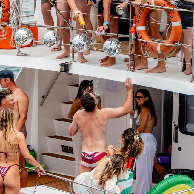 A group of people enjoying a lively yacht party. They are in swimwear, with some holding drinks and others laughing. The yacht has orange life rings and shiny disco balls as decorations, creating a festive vibe akin to an exclusive social club on the water.