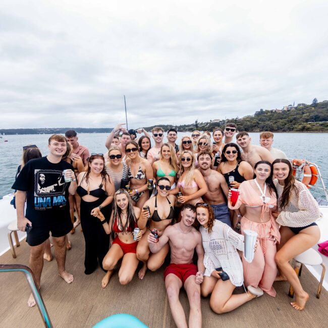 A large group of people in swimwear are posing for a photo on the yacht deck. They are smiling and holding drinks, with a scenic view of water and a wooded coastline in the background. The sky is overcast, adding to the cozy vibe of this social club gathering at sea.