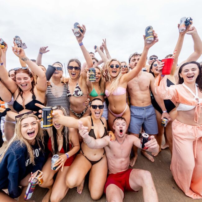 A lively social club gathers on a yacht, wearing swimwear and holding drinks while cheering against an ocean backdrop with a cloudy sky. Some sport sunglasses as they revel in the festive atmosphere, their excitement palpable amidst the waves.