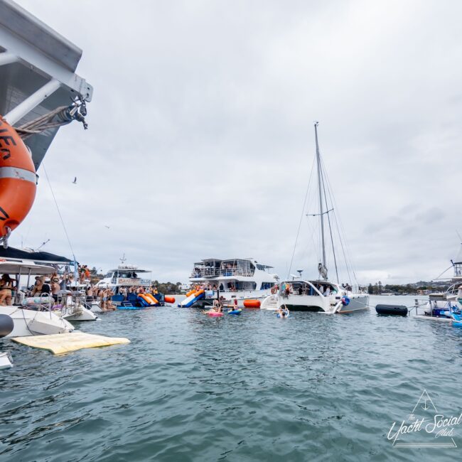 A group of boats gathered on the water under a cloudy sky, reminiscent of a yacht club gathering. People are socializing and enjoying the scene, with some on inflatables near the boats. A large yacht is prominent in the center, while a buoy and life preserver are visible on the left.