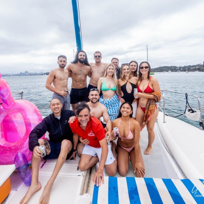 A group of people in swimwear are smiling and posing on a yacht. The scene, reminiscent of a vibrant social club gathering, features inflatable floats, cloudy skies, and stunning waterfront views.