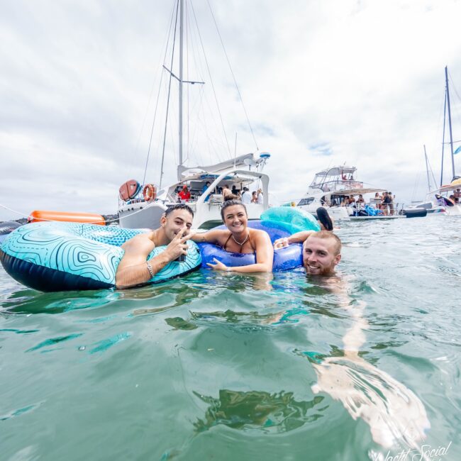 Three people enjoy the water on inflatable floats near sailboats, with a majestic yacht in the distance. The sky is overcast, and several other boats and fellow members of their social club are in the background, adding to the lively scene.