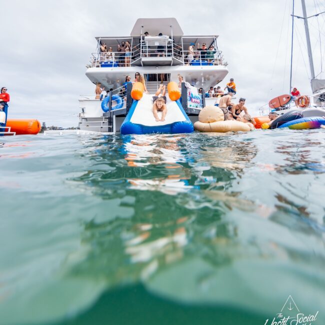 People on a yacht enjoying a party, with several individuals on inflatable slides and floats in the water. The scene suggests a festive atmosphere reminiscent of an exclusive social club on the open seas, with a mix of sunny and cloudy weather adding to the charm.