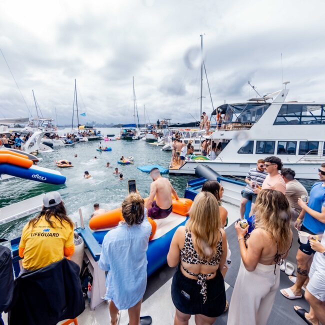 A lively scene of people in swimsuits enjoying a party on yachts anchored in a bay. The sky is cloudy, and several inflatable floats can be seen in the water. Vibrant social club atmosphere with music and mingling.