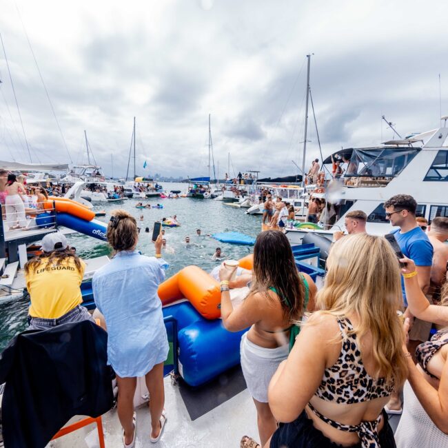 People are enjoying a vibrant Social Club yacht party on the water, surrounded by various boats and inflatables. Some are swimming, while others socialize on deck. The sky is cloudy, and the atmosphere is lively and festive.