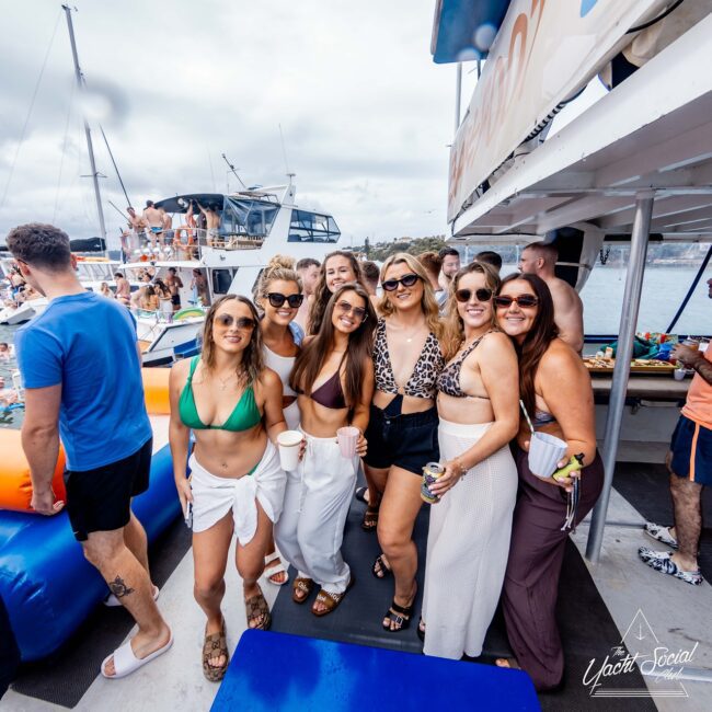 A group of smiling people poses for a photo on a yacht. They are in swimwear, holding drinks, with a festive atmosphere. Other people and boats are in the background, indicating a lively social club gathering on the water.