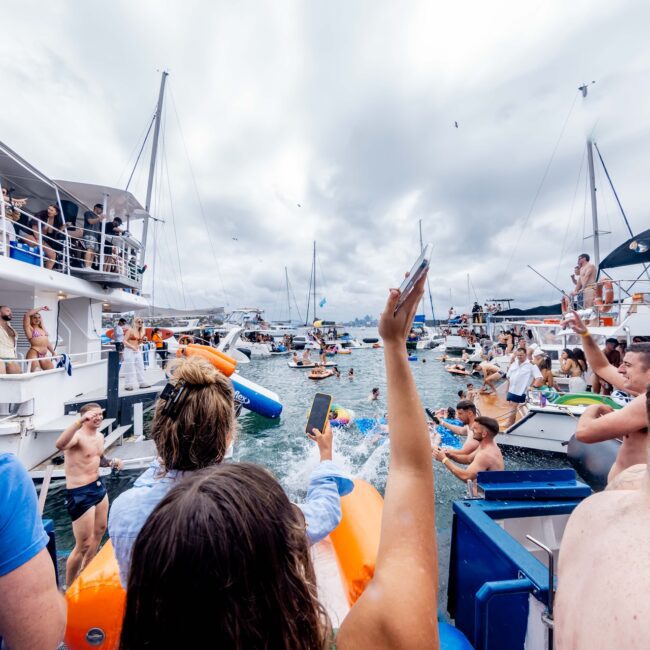 A lively yacht party unfolds on a cloudy day, where members of the social club bask in the festive atmosphere. Guests mingle on boats or in the water, drinks in hand and smiles wide, capturing memories. Music and laughter fill the air amid a backdrop of elegantly anchored yachts.