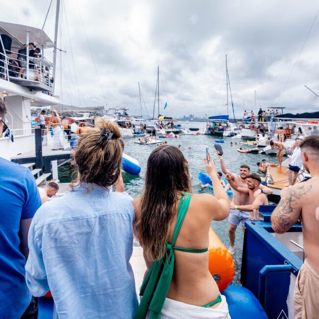 People gathered on a yacht for a lively party, part of an exclusive social club event, with others swimming and socializing in the ocean. Boats are scattered in the background under a cloudy sky. The scene captures a vibrant, nautical celebration.