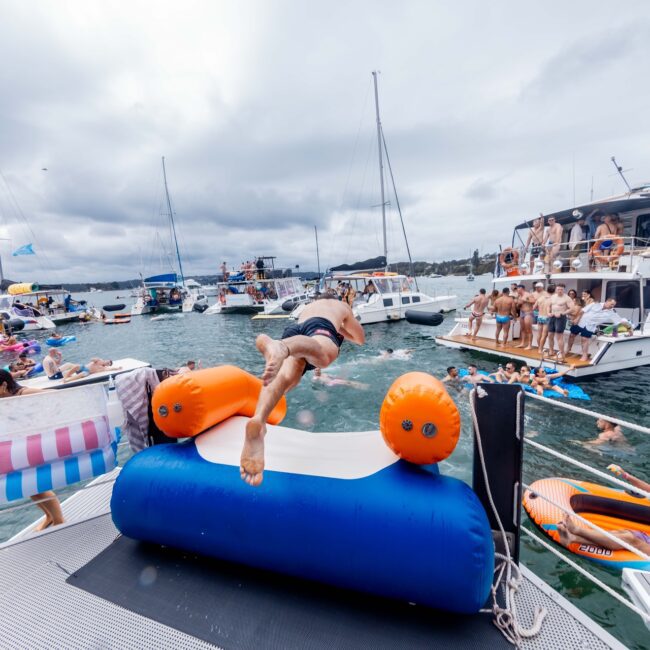 A person is diving into the water from an inflatable toy on a yacht. Surrounding boats are filled with people reveling in a Yacht Social Club atmosphere. Some are swimming, others lounging on inflatables, all under a cloudy sky.