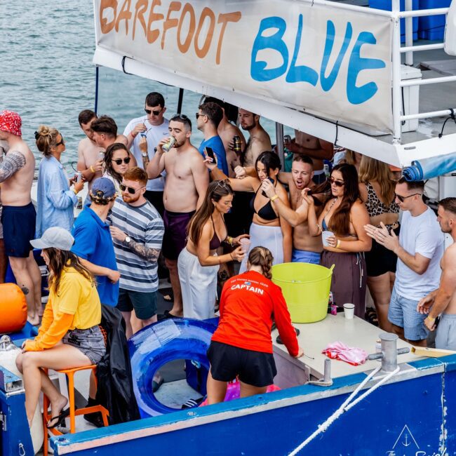 A lively group of people from a local social club enjoys a festive party on the yacht "Barefoot Blue." Mingling with drinks in hand and some in swimwear, they soak up the relaxed atmosphere. A person in a red captain's shirt stands near an ice bucket, embodying the carefree spirit of the day.