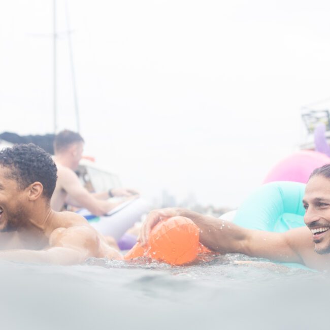 Two people smile and play in the water, surrounded by colorful inflatable tubes. Boats and more people appear in the blurred background, suggesting a joyful, relaxed atmosphere on a sunny day.