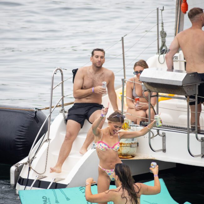 A group of people enjoying a day on a boat. Two women in swimsuits dance on a floating pad in the water, holding drinks. Others are seated on the boat, watching and relaxing. The atmosphere is festive and lively.