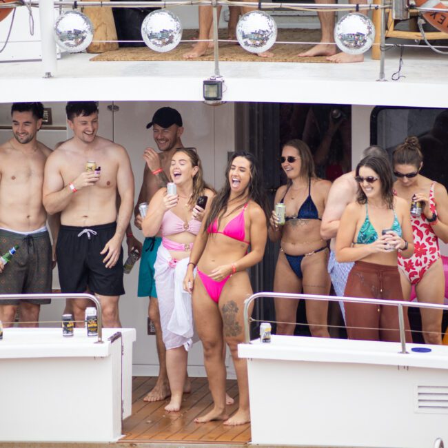 A group of people in swimwear is enjoying themselves on a boat. They are smiling, holding drinks, and appear to be having a good time. Various colorful pool floats are visible in the background.