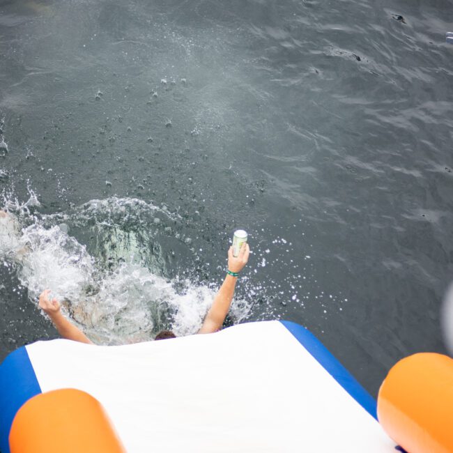 Person sliding into a body of water from an inflatable slide, splashing water around. Their arms are raised, holding a small object. An orange inflatable object is visible nearby on the water surface.