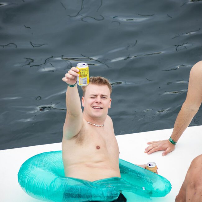 A man in a green inflatable inner tube on a floating platform holds up a can while smiling. He is accompanied by two other men, all shirtless, enjoying a day on the water. The background shows dark, rippling water.