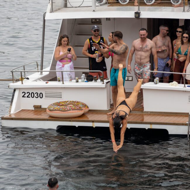 A group of people on a boat enjoying a party. A woman in a black bikini is diving into the water, while others watch and take photos. The boat has a donut float and various people are holding drinks. The atmosphere appears festive and lively.