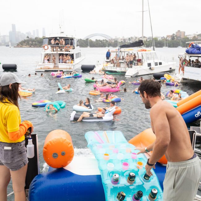 A lively scene on the water with people on colorful inflatables, surrounded by boats. One person stands on a giant inflatable with a cup-holder pattern, while others relax and socialize. The skyline and a bridge are visible in the background.