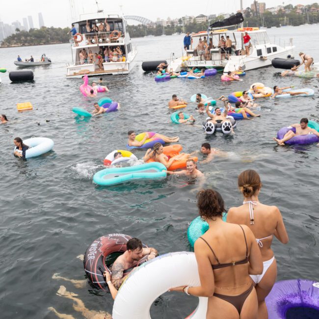 A group of people in swimsuits enjoy a party on the water, using colorful inflatable rings and floats. Several boats are anchored nearby, and a city skyline is visible in the distance on an overcast day.