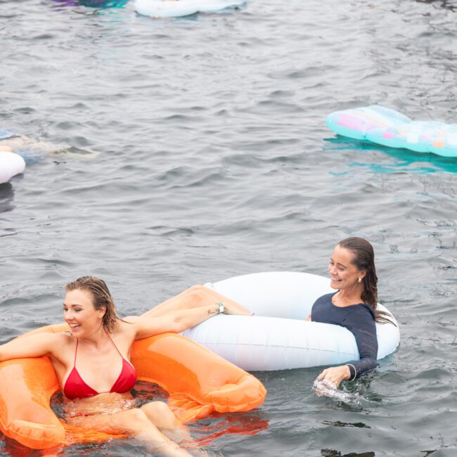 People enjoying a swim in the water, lounging on inflatable rafts. One person in a red bikini leans on an orange float, while another wears a dark shirt and sits on a white and blue float. Other colorful inflatables can be seen in the background.