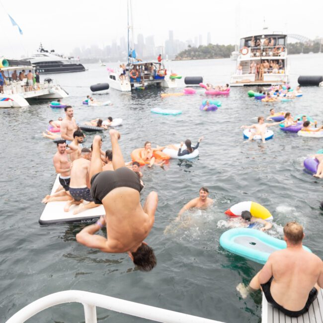 People are enjoying a party on the water, surrounded by boats and inflatables. A person is mid-flip, jumping into the water. Various people are lounging and swimming, creating a lively atmosphere. The background shows a cityscape and other boats.