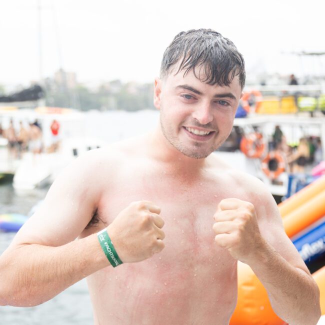 A man with wet hair stands in front of a harbor, smiling and raising his fists. People and inflatable toys are visible in the background on boats. He wears a wristband and no shirt. It's a bright, cloudy day.