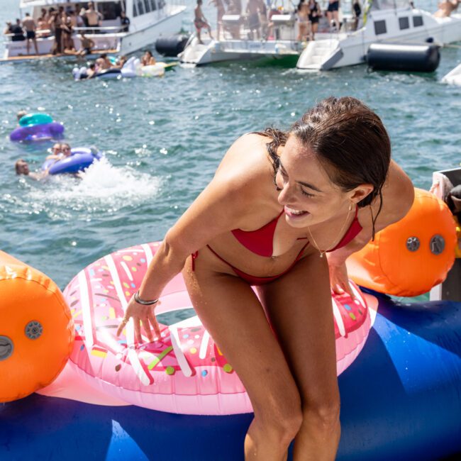 A woman in a red bikini smiles while sitting on a colorful inflatable toy on the water. Multiple boats and people are in the background, enjoying a sunny day.