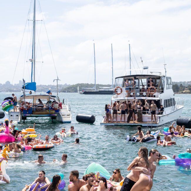 A lively scene of people enjoying a sunny day on the water, with groups on colorful inflatables near two boats. The foreground shows people socializing on a floating platform. Sailboats and city skyline are visible in the background.