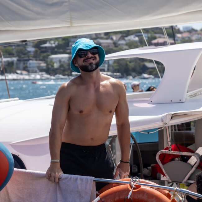 Two men are standing on a boat under the sun. One is wearing a blue bucket hat and sunglasses, smiling. The other is in the background. The boat is on a body of water with trees and buildings in the distance.