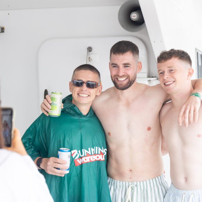 Three smiling men pose for a photo on a boat. One man wears a green poncho and sunglasses, holding a drink, while the other two are shirtless. A person in the foreground takes their picture.