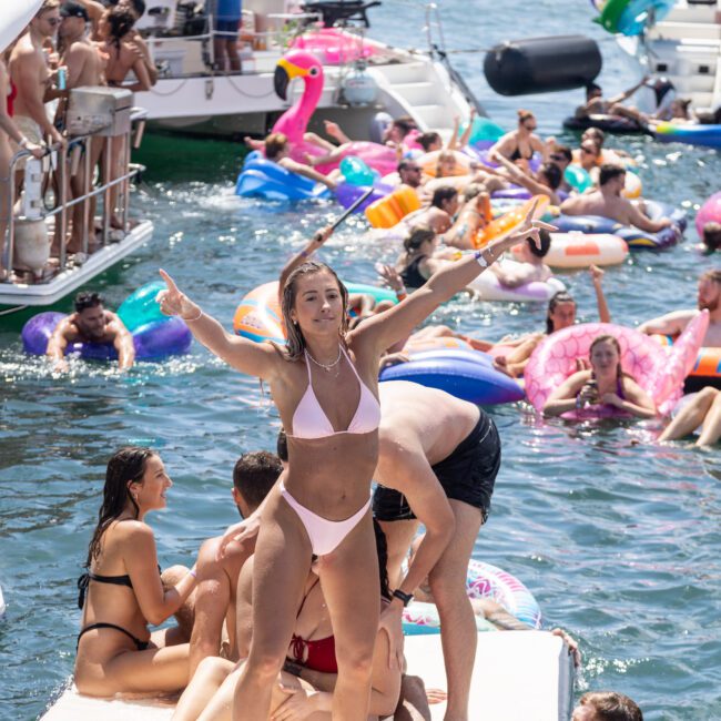 A woman in a white bikini dances on a floating platform surrounded by people and inflatable floats in the water. Boats are anchored nearby under a sunny sky, with a cityscape visible in the background.