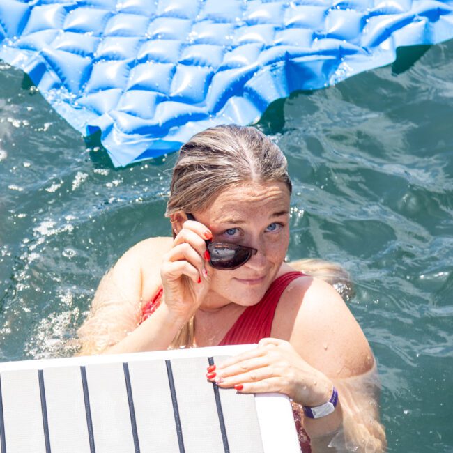 A person with light hair, wearing a red swimsuit and sunglasses, is in the water next to a blue floating mat. They hold onto the edge of a platform, partially submerged in the clear water under bright sunlight.