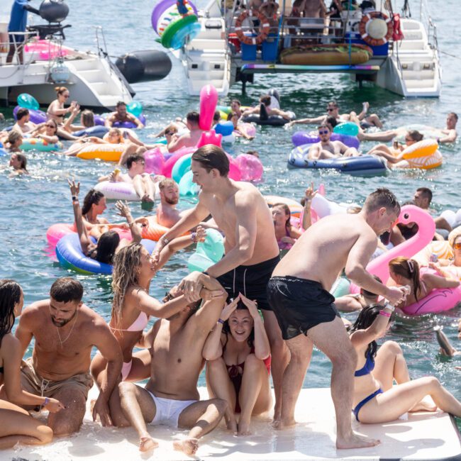 A lively scene of people enjoying a sunny day on the water. They are on floats and boats, laughing and playing. The image is colorful, with inflatables in shapes like flamingos and unicorns, and a crowded boat in the background.