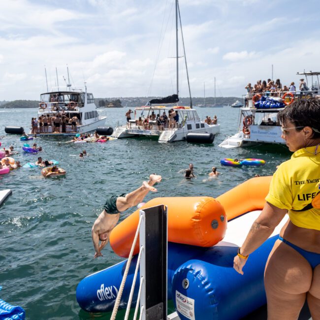 People enjoy a sunny day on the water, swimming and lounging on floats near several boats. A lifeguard in a yellow shirt watches the scene while someone dives off a platform.