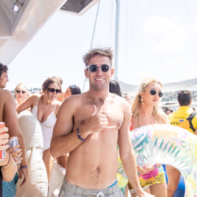 A group of people enjoying a sunny day on a boat. A shirtless man in sunglasses is smiling and making a hand gesture. Others around him hold drinks and inflatable toys, with a bright sky in the background.