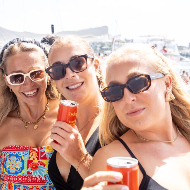 Three women wearing sunglasses smile and hold drinks on a boat under a sunny sky. They are surrounded by other people and boats in the background, enjoying a festive atmosphere.