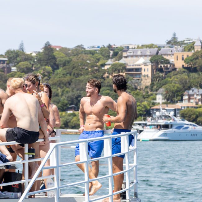 A group of people in swimwear socializing on a boat with a scenic background of a coastal town, trees, and docked yachts. The water is calm and the weather is sunny.