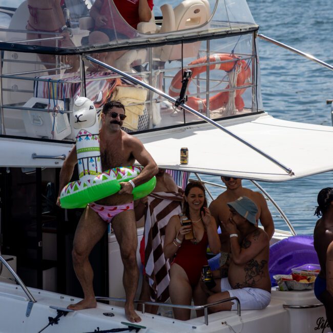 A man wearing pink swim briefs stands on a boat holding a llama inflatable. He is surrounded by a group of people enjoying themselves on the deck. The boat is on a body of water under clear skies.