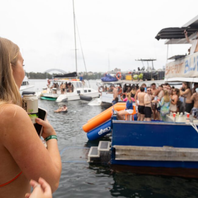 A woman in a swimsuit holds a drink on a boat's deck, looking at a lively party on a nearby boat. The scene includes people in swimwear, inflatable toys, and boats floating on the water under an overcast sky.