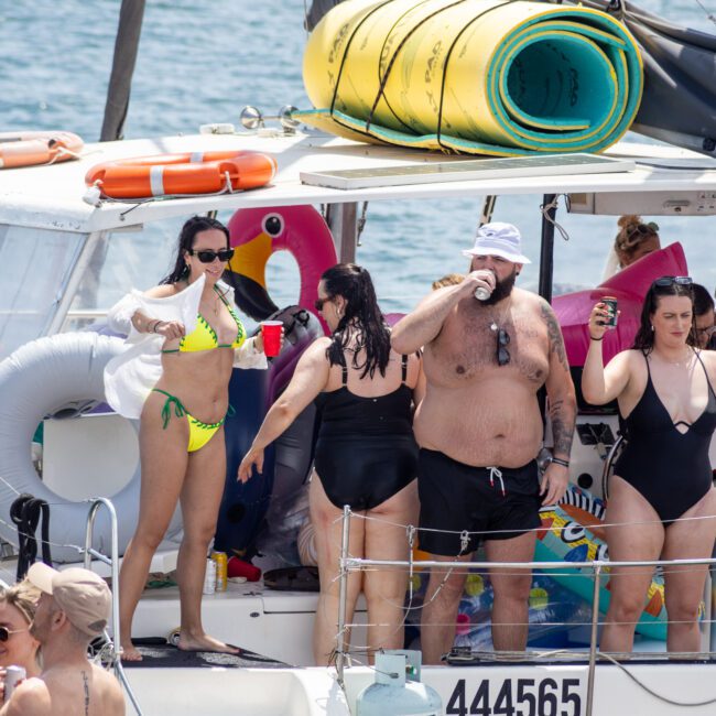 A group of people in swimsuits enjoying a sunny day on a sailboat. They are laughing, holding drinks, and surrounded by inflatable pool floats and beach gear. The boat is on a body of water, with more people visible in the background.