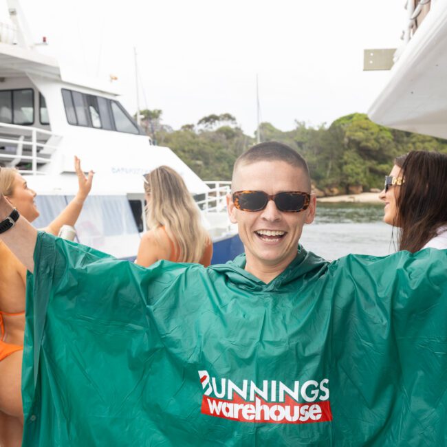 A person smiles widely, wearing sunglasses and a green "Bunnings Warehouse" poncho while holding a canned drink. They're on a boat with other people, and another boat is in the background near trees.