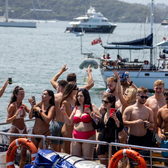 A group of people enjoying a party on a boat. The scene includes men and women in swimwear, holding drinks, and dancing. The background shows a calm body of water with several boats and greenery on the shore.