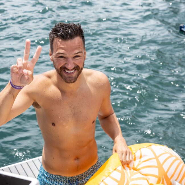 A man in swim trunks stands on a dock, smiling and holding up a peace sign. He has short dark hair and a beard. An inflatable float is beside him, and the water is sparkling in the background.