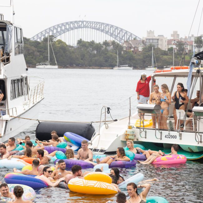 People enjoying a boat party on a river with inflatable floaties. Two boats are docked nearby. The iconic arch of a large bridge is visible in the background, along with city buildings and trees.